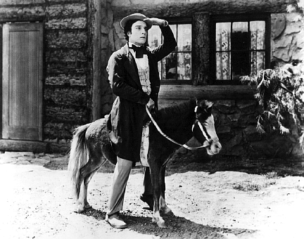 Buster Keaton riding on a minauture horse in front of a log cabin.